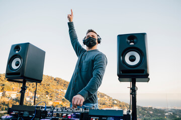 Professional DJ with protective face mask playing music raising his hand to cheer up the crowd of...