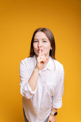 Portrait of smiling beautiful girl in white blouse showing silence with hand, on yellow isolated background