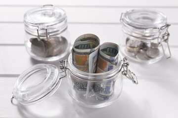 Glass jars with coins and cash on a white wooden background.