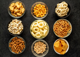 Assorted chips, popcorn, onion rings, croutons, cheese balls, biscuits in bowls. unhealthy food for figure, heart, skin, teeth. An assortment of fast carbohydrates. Junk food on a stone background
