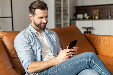Attractive millennial young bearded man with a pleasant smile chilling and sitting on the couch, chatting with friend in social media on mobile phone, dating, flirting, spending leisure time online