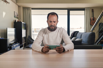 middle-aged bearded man looking at his cell phone with a worried face sitting around a table in the middle of his loft apartment