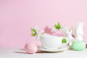 White mug with tea or coffee near pink and green Easter eggs, rabbits and flowers, on a concrete white table, on a pink background, with hard shadows. Easter breakfast concept.