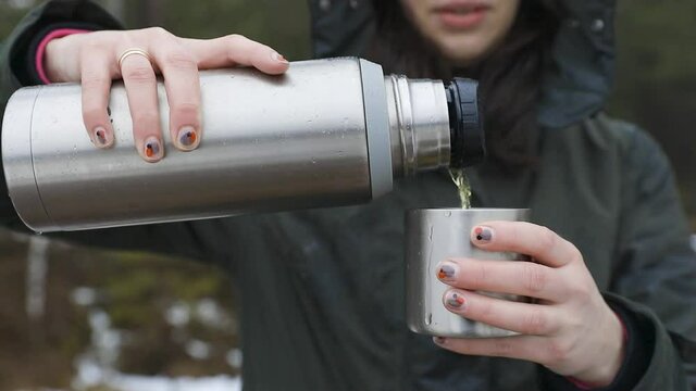 Young beautifull women pouring beverage from termos to cup in a cold raini day. Healthy living and hiking.