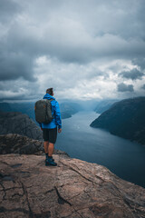 A man with a blue jacket, shorts, hiking boots, and backpack standing on the top of the cliff Preikestolen and overlooking a fjord in Norway. Moody landscape and sky.