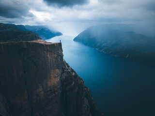 A man with a blue jacket, shorts, hiking boots, and backpack standing on the top of the cliff Preikestolen and overlooking a fjord in Norway. Moody landscape and sky.