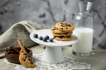 Horizontal capture of chocolate chip cookies lined upwards with bottle of milk, blueberries, cinnamon bark in wooden plate and lace ground in frame
