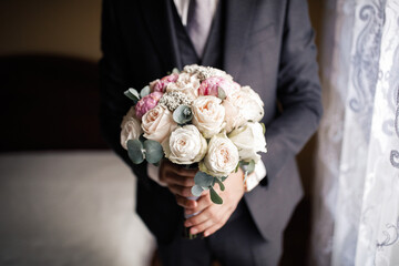 Man groom in a beautiful stylish classic suit holds a wedding bouquet of white and pink roses standing at the window waiting for the bride. Hands without a face.