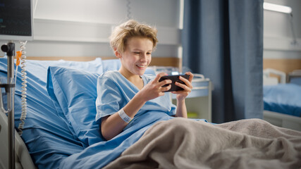 Hospital Ward: Handsome Young Boy Resting in Bed, Uses Smartphone, Plays Online Video Games on Internet with Mobile Phone. Happy Patient Recovering after Sickness