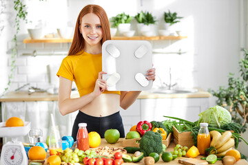 Fitness redhead woman with weight scale standing behind table with fruit and vegetables in kitchen