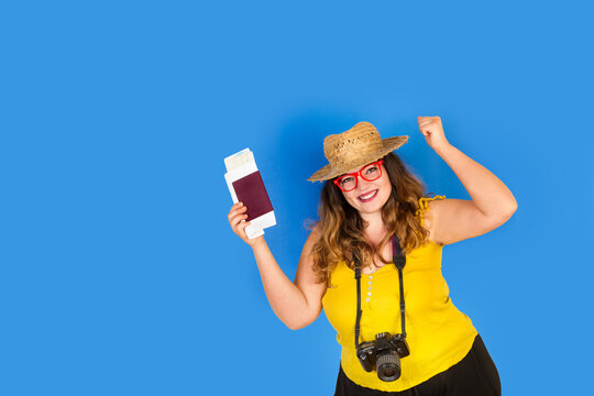 Mujer joven sonriente con pasaporte y boletos de tarjeta de embarque en las manos y con cámara de fotos sobre un fondo azul liso y aislado. Vista de frente. Copy space