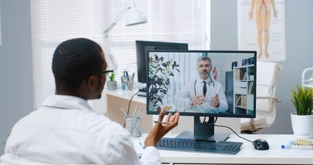 Over shoulder view of African American male specialist talks on online video call with Caucasian middle-aged doctor on computer sitting in office experienced physician consults nurse on web conference - obrazy, fototapety, plakaty
