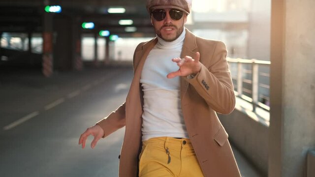 Cheerful and happy young man with beard actively dancing while walking down the street on sunset background. He's wearing a brown jacket. Urban concrete park next to business center. Sunny.