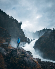 A man with a blue jacket standing by the waterfall Låtefossen in Norway. Moody weather.
