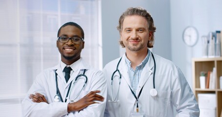 Portrait of mixed-races happy male doctors colleagues in white coats standing in cabinet in hospital, looking at camera and smiling in good mood. Professional physicians, healthcare workers, hospital