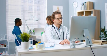 Portrait of happy bearded Caucasian middle-aged male doctor working in hospital cabinet typing on computer while female physician talking with African American patient on background