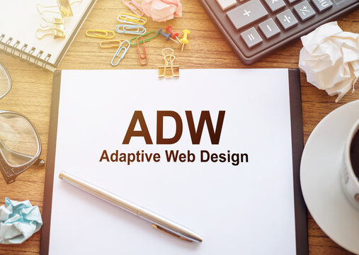 On a wooden table there is an office sheet of paper with the text ADW - Adaptive Web Design. Business workspace with calculator, glasses, pen, crumpled paper and cup of coffee.