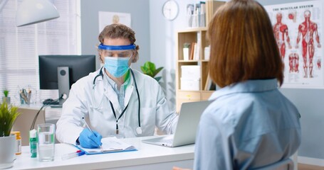 Portrait of Caucasian male general practitioner wearing medical mask and protective face shield having consultation in hospital with female patient writing down symptoms, coronavirus, healthcare