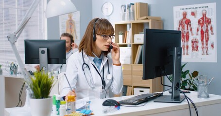 Portrait of beautiful young female Caucasian doctor in headset speaking on video call on computer showing X-ray scan to patient explaining treatment while sitting in hospital at desk, clinic concept