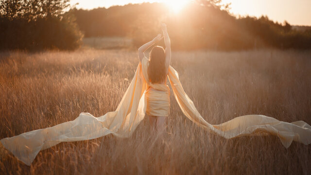 A young girl with long red hair in summer in a long yellow dress wrapped around her open body and arms is standing in a field looking at the sunset and horizon. Beautiful woman stands alone in nature