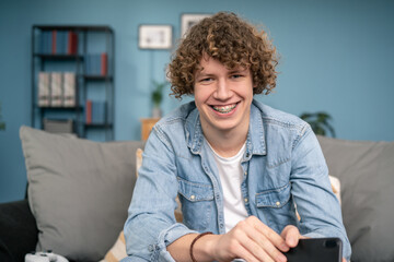 Portrait of Hendsome Young man with Light Brown Curly Hair Wearing blue shirt Looking Up to the Camera and Smiling Charmingly. Successful man Resting in Bright Living Room.