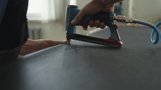 A professional worker attaches fabric to a wooden frame with a pneumatic stapler in an upholstered furniture workshop