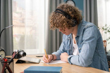 Fototapeta na wymiar Focused american teenage boy wearing headphones writing notes study with laptop and books, serious white man high school teen student listening audio course or music while doing homework