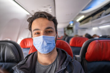 handsome brown-haired man with mask smiles while sitting on a plane seat in quarantine days with copy space