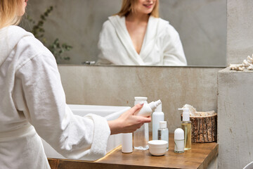pretty model in bathrobe using dispenser of white bottle with cream mask, standing in front of mirror in bathroom