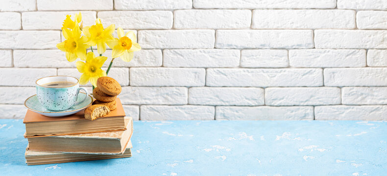Spring flowers daffodils bouquet, coffee cup, books, cookies on blue table over white brick background. Reading and breakfast, cozy home interior. Spring, hygge, spring holidays, read books concept