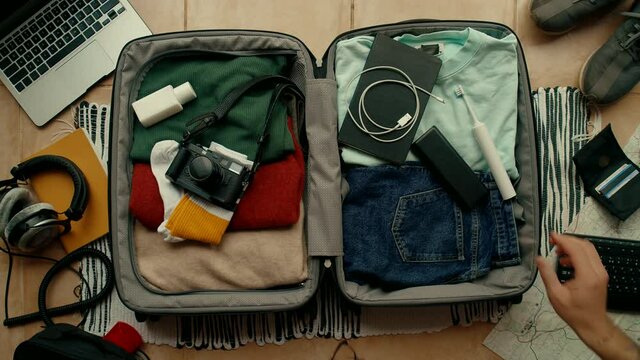 Urban traveller, man with arm tattoos packs his suitcase with travel essentials, electric toothbrush, laptop, power bank and photo camera. Man puts hydration water bottle flask in bag