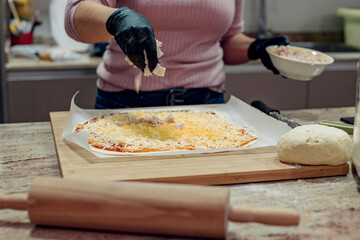 Detalle de una mujer poniendo pequeños trozos de jamón en una pizza casera con salsa de tomate y mozzarella. Lleva guantes. Encimera de cocina con un rodillo y una tabla de cortar de madera