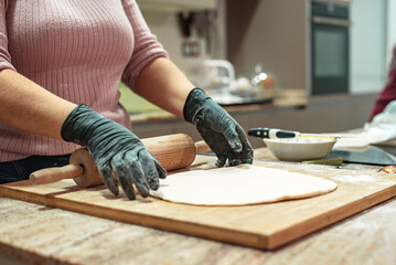 Detalle de las manos de una mujer con guantes amasando una masa de pizza casera con un rodillo sobre una encimera de cocina de madera. Concepto de hacer pan en casa