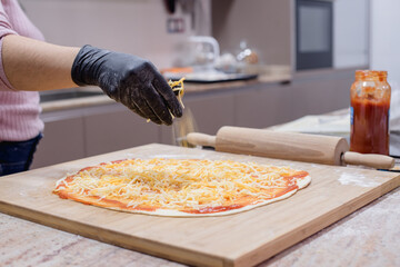Detalle de una mujer echando queso mozzarella en la masa de una pizza casera con salsa de tomate mientras usa guantes. Mostrador de cocina con un rodillo y una tabla de cortar de madera