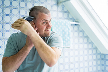 A man of forty years old staying in a bathroom and shaving himself with electronic razor, taking...