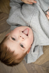little boy child showing tongue emotions lying on the brown wooden floor upside down in grey clothes