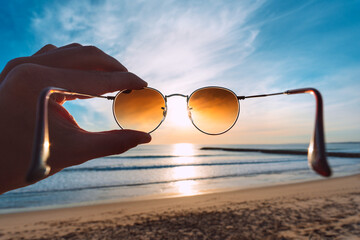 Hand holding stylish round sunglasses with brown lenses at sunset. Putting on sunglasses at sunny...