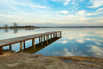 Long wooden jetty towards a calm lake, reflection of the clouds in the water