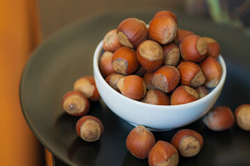 Heap, pile of brown nuts, hazelnuts lies in a small white plate and a black plate on table on blurry warm background. Macro
