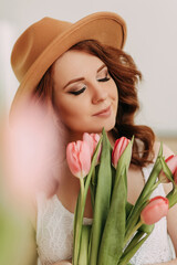 A close up portrait of a beautiful tender sensual elegant red haired young woman in a fashionable hat holding and smelling a bouquet of spring flowers tulips enjoying the fragrance