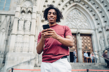 Happy travel blogger with curly hair using cellphone gadget for social networking during journey vacations for visiting historic city, smiling hipster guy connecting to 4g internet for browsing