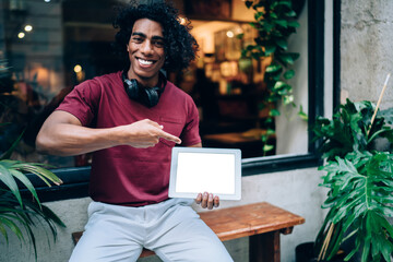 Portrait of joyful millennial user with bluetooth earphones pointing on blank digital gadget and...