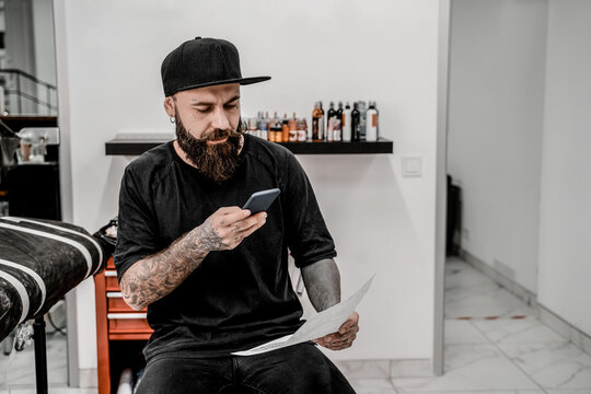 Young male tattoo artist with beard taking picture of sketch sitting on couch in workshop place.