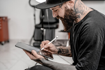 Young man tattoo artist with beard holding pencil and sketch looking positive and happy standing and smiling in workshop place.