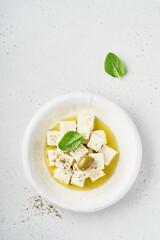Feta cheese cubes with rosemary, olives and olive oil sauce in white bowl on light gray background. Traditional Greek homemade cheese. Selective focus.
