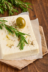 Cheese feta with rosemary, herbs, olives and olive oil on wooden cutting board on old wooden background. Traditional Greek homemade cheese. Selective focus.