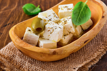 Feta cheese cubes with rosemary, olives and olive oil sauce in bowl on old brown wooden background. Traditional Greek homemade cheese. Selective focus.