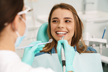 Smiling woman sitting in medical chair while dentist fixing her teeth - obrazy, fototapety, plakaty