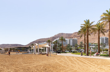 View of the beach on the Ein Bokek embankment on the coast of the Dead Sea, tourist hotels and sandy mountains in the background, in Israel
