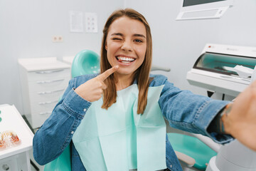 Joyful woman winking and pointing finger at her smile while taking selfie